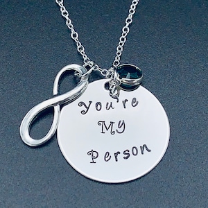May include: Silver necklace with an infinity symbol charm and a round pendant that says "You're My Person". A small green gemstone is attached to the chain.