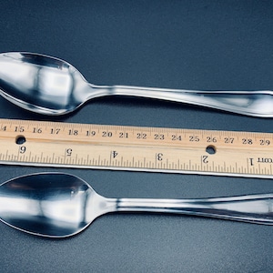 May include: Two silver spoons with rounded bowls and slightly curved handles. The spoons are placed on a black surface next to a wooden ruler marked with inches.