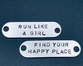 Puede incluir: Dos etiquetas de metal plateado con agujeros. La etiqueta superior dice "Run Like A Girl". La etiqueta inferior dice "Find Your Happy Place".
