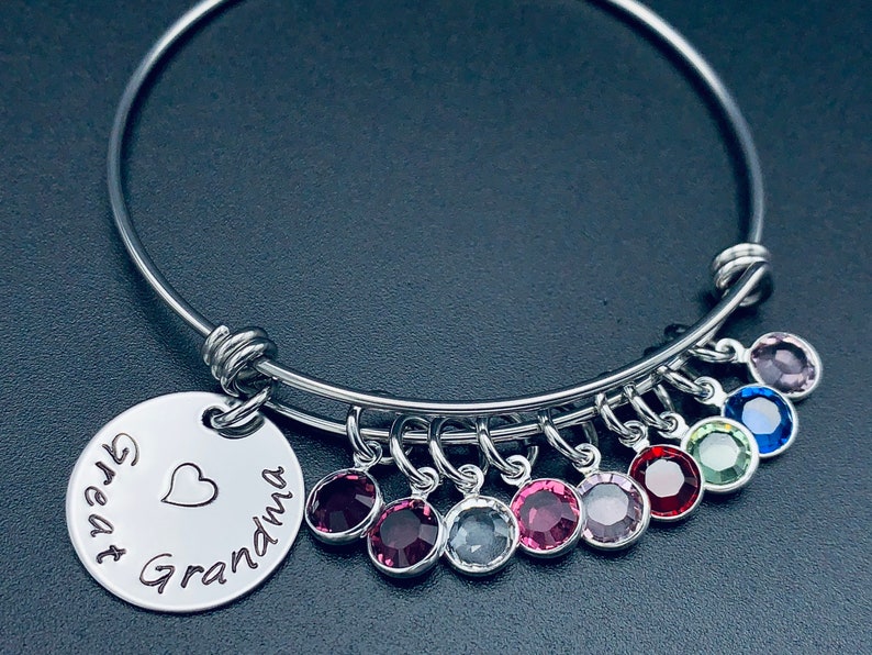 May include: A silver bangle bracelet with a charm that reads "Great Grandma" with a heart. The bracelet has eight dangling charms with colourful crystals.
