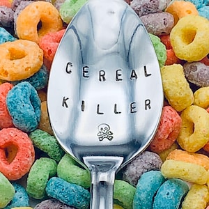 May include: A silver spoon with the words "Cereal Killer" engraved on it, surrounded by colorful cereal rings.