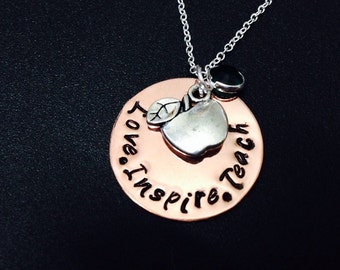 Op de afbeelding: Een koperen ketting met een zilveren appelbedel en de woorden "Love Inspire Teach" gegraveerd op de hanger. Een donkergroene edelsteen hangt aan de ketting.