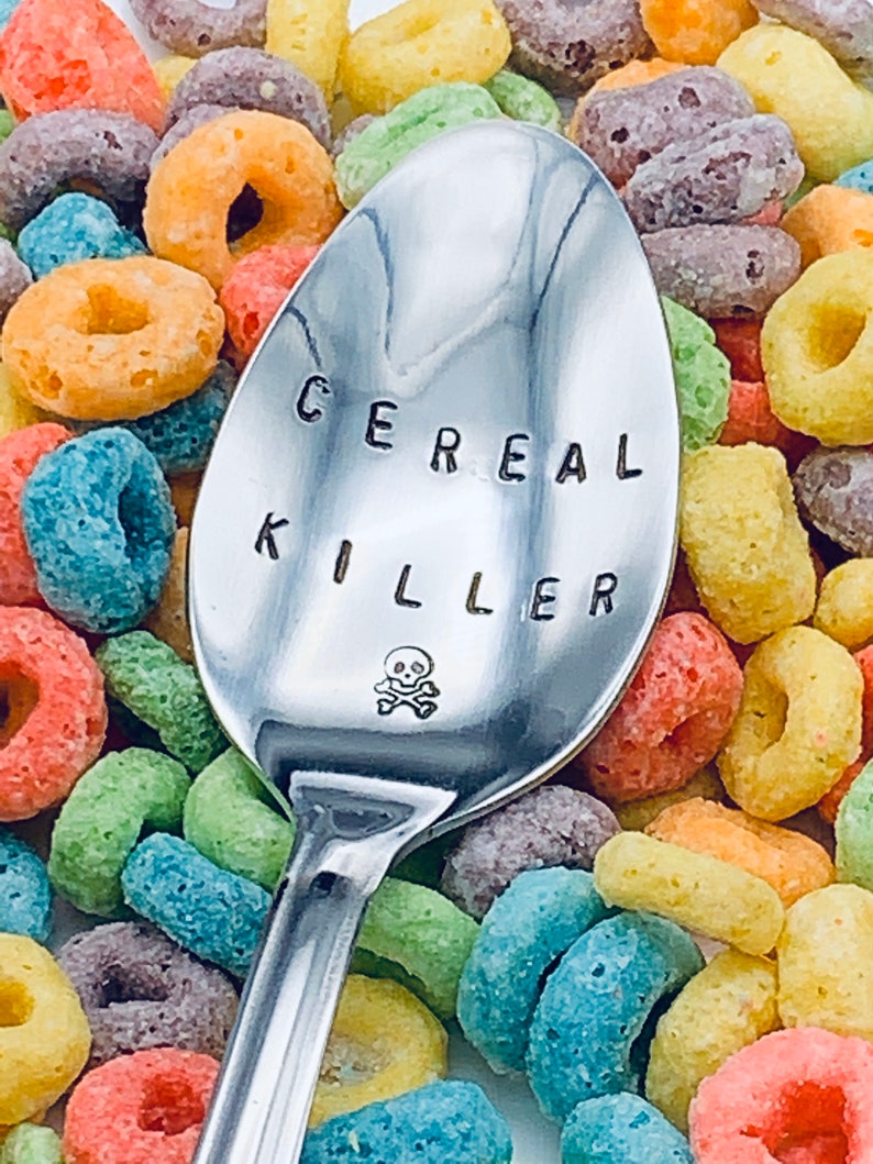 May include: A silver spoon with the words "Cereal Killer" engraved on it, with a skull and crossbones symbol below the text. The spoon is surrounded by colorful cereal rings.
