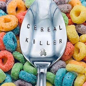 May include: A silver spoon with the words "Cereal Killer" engraved on it, with a skull and crossbones symbol below the text. The spoon is surrounded by colorful cereal rings.