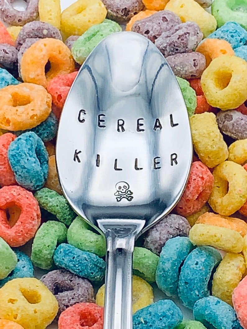 May include: A silver spoon with the words "Cereal Killer" engraved on it, with a skull and crossbones symbol below the text. The spoon is surrounded by colorful cereal rings.