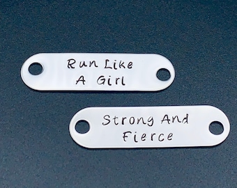 May include: Two silver oval-shaped tags with holes for attaching to a keychain or bag. The top tag reads "Run Like A Girl". The bottom tag reads "Strong And Fierce".