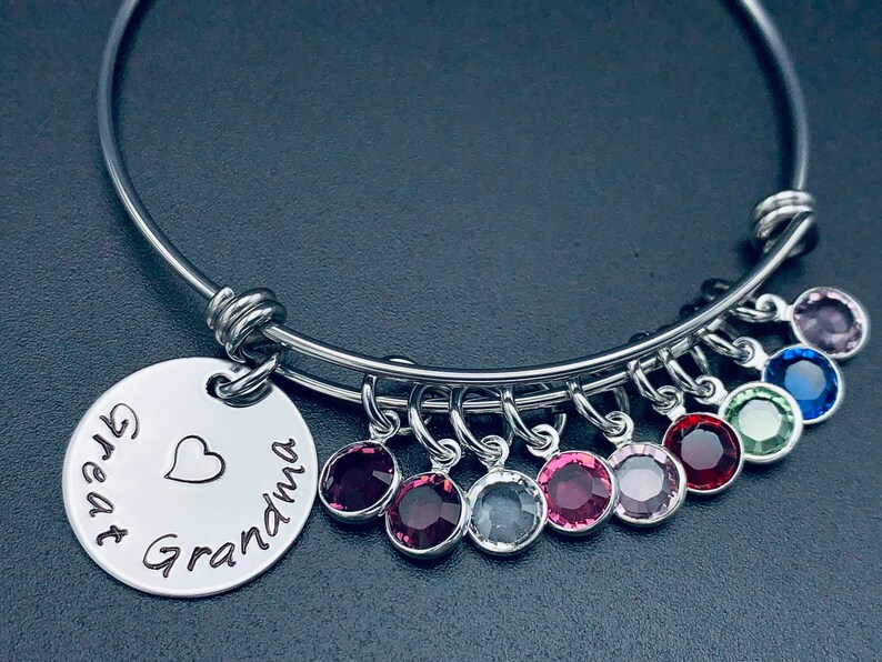 May include: A silver bangle bracelet with a charm that reads "Great Grandma" with a heart. The bracelet has multiple charms with colourful crystals.