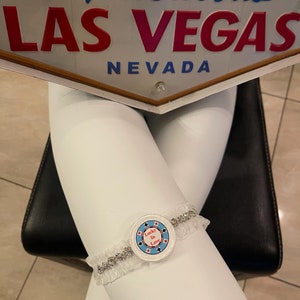 Op de afbeelding: Een witte kousenband met een zilveren rand en een ronde blauwe en witte pokerchip-hanger met de tekst "Lucky in Love".