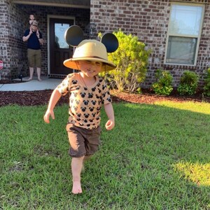 May include: A young child wearing a brown and tan safari hat with Mickey Mouse ears, a brown and tan short-sleeved shirt with a Mickey Mouse print, and brown shorts. The child is standing on a green lawn.