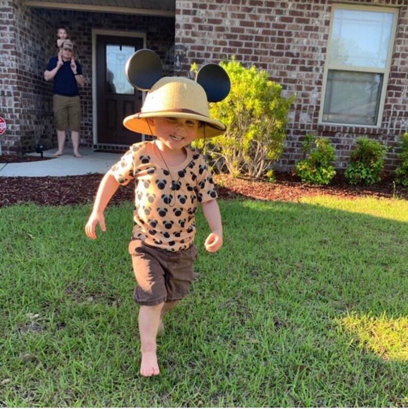 May include: A young child wearing a brown and tan safari hat with Mickey Mouse ears, a brown and tan short-sleeved shirt with a Mickey Mouse print, and brown shorts. The child is standing on a green lawn.