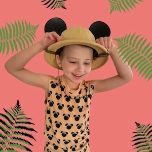 May include: A young person wearing a tan safari hat with Mickey Mouse ears, a yellow sleeveless shirt with a Mickey Mouse pattern, and a big smile. The background is pink with green ferns.