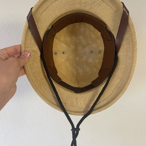 May include: A tan straw hat with a brown leather strap and a black chin strap. The hat has a mesh lining and a wide brim.