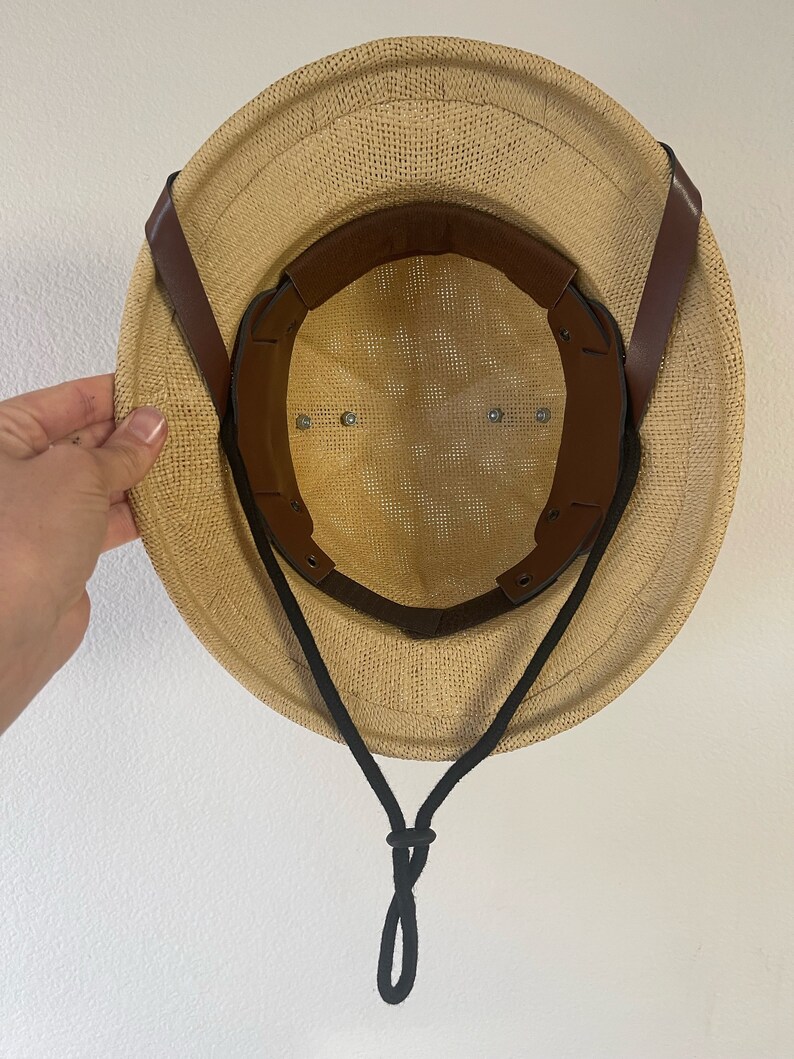 May include: A tan straw hat with a brown leather strap and a black chin strap. The hat has a mesh lining and a wide brim.
