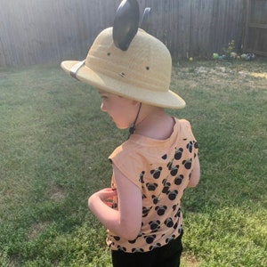 May include: A child wearing a tan safari hat with black mouse ears and a peach colored shirt with a black Mickey Mouse pattern.