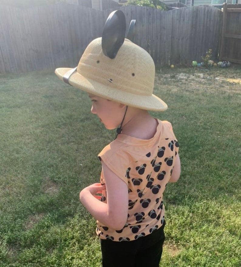 May include: A child wearing a tan safari hat with black mouse ears and a peach colored shirt with a black Mickey Mouse pattern.