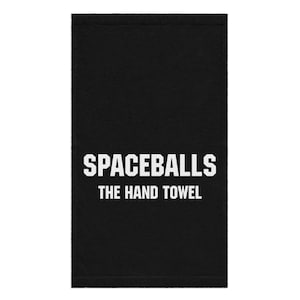 May include: Black hand towel with the text "SPACEBALLS THE HAND TOWEL" in white block letters. The towel is rectangular and appears to be made of a soft, absorbent material. The design is simple and bold.