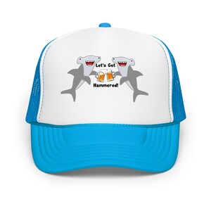 Könnte beinhalten: Blau-weiße Trucker-Cap. Die Vorderseite ist weiß, mit einem blauen Schirm und Mesh-Rückseite. Sie zeigt einen Cartoon von zwei Hammerhaien, die Bierkrüge anstoßen, mit dem Text "Let's Get Hammered!"