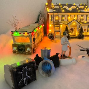 Könnte beinhalten: Eine Miniatur-Weihnachtsdorf-Szene mit einem Haus, einem Wohnmobil und anderen dekorativen Elementen. Das Haus ist mit Lichterketten beleuchtet, und eine Leiter lehnt daran. Das Wohnmobil hat grüne und rote Lichter. Ein kleiner Stuhl mit einer Katzen-Silhouette befindet sich im Vordergrund.