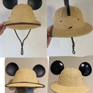 May include: A tan safari hat with black Mickey Mouse ears attached. The hat has a brown leather band around the brim and a black chin strap.