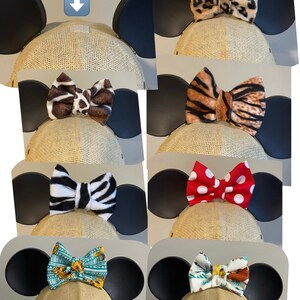 May include: Five different bow designs for a Mickey Mouse ear hat. The bows are made of fabric with various patterns, including animal print, polka dots, and a cartoon design. The text "Permanent Velcro" is visible in the top left corner of the image.