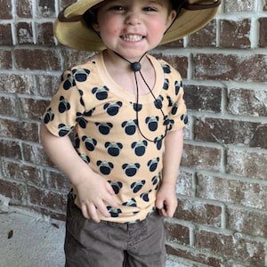 May include: A young child wearing a tan safari hat with black Mickey Mouse ears attached. The hat has a black chin strap with a toggle. The child is wearing a tan t-shirt with a black and white Mickey Mouse pattern. The child is also wearing brown shorts. This is the old chin strap. The new one included is thicker and pictured in the other pics. Strap toggle could be a choking hazard. Please remove if needed.