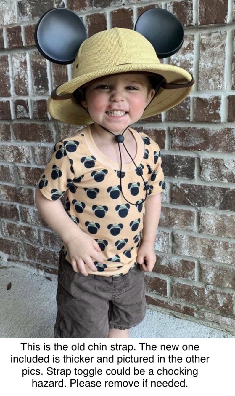 May include: A young child wearing a tan safari hat with black Mickey Mouse ears attached. The hat has a black chin strap with a toggle. The child is wearing a tan t-shirt with a black and white Mickey Mouse pattern. The child is also wearing brown shorts. This is the old chin strap. The new one included is thicker and pictured in the other pics. Strap toggle could be a choking hazard. Please remove if needed.