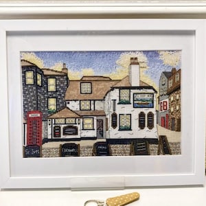 May include: Framed cross-stitch art showing a detailed street scene with a pub, telephone box, and buildings. The artwork has a white frame and is displayed on a white surface. The scene includes the text "The Sloop Inn" and "St Ives Cornwall".