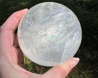 May include: A large, clear, iridescent selenite sphere. The sphere is smooth and polished, with a slight sheen. The sphere is held in a hand, and the background is blurred.