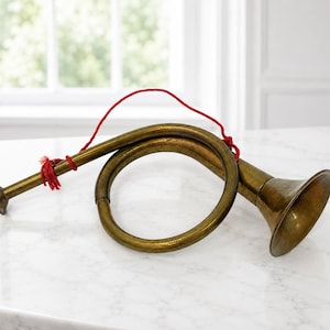 May include: A curved brass hunting horn with a polished metallic finish and a wide, flared bell. The instrument features a thin, bright red string tied around the narrow neck and the circular body, providing a contrast to the aged brass surface.