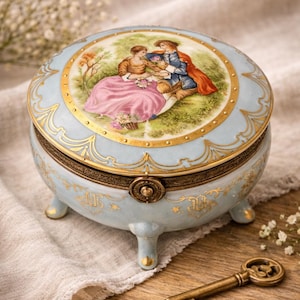 May include: A light blue, round trinket box with a lid featuring a painted scene of a couple in a garden. The box has gold accents, a decorative clasp, and three small feet. A vintage-style key rests nearby.