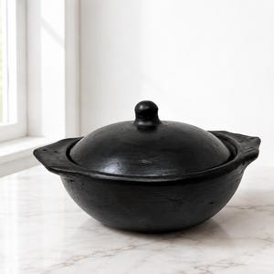 May include: A matte black, round, lidded cooking pot with two side handles. The vessel features a smooth, dark finish and a domed lid with a small central knob. The design is simple and functional, showcasing a deep, circular basin shape.
