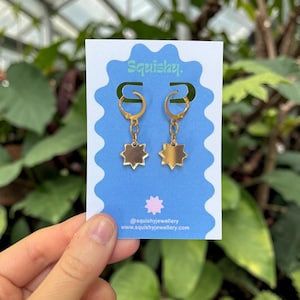 May include: Gold hoop earrings with star-shaped pendants. The earrings are displayed on a blue card with the text "Squishy." and social media details. Green foliage forms the backdrop.