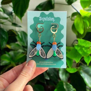 May include: Fly-shaped earrings with gold hoops. Each earring has a blue body, orange head, and green accents. The wings are transparent. The earrings are displayed on a teal card with the word "Squishy." The brand's website is also visible.