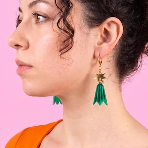 May include: A pair of gold-toned hoop earrings featuring a dangling star charm and a translucent green, multi-faceted, bell-shaped pendant. The earrings are worn on the earlobes, showcasing the metallic star and the vibrant green geometric drop design.