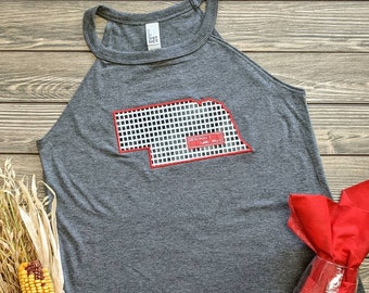 May include: Gray tank top with a red outline of the state of Nebraska. The state is filled with a checkered pattern. The text "Nebraska" is written in red inside the state outline.