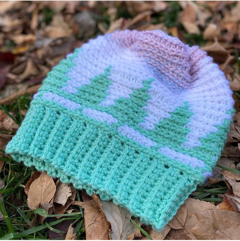 Pu&ograve; includere: Un cappello all'uncinetto con un design sfumato in verde, bianco e rosa. Il cappello presenta un motivo di alberi verdi su uno sfondo bianco.