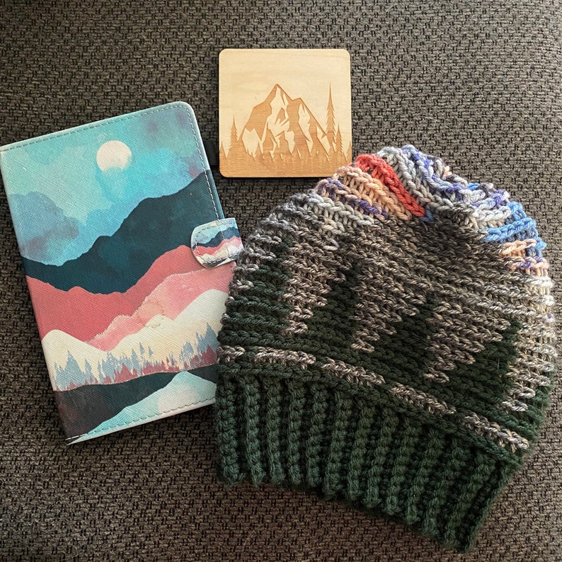 Pu&ograve; includere: Un berretto lavorato a maglia di colore verde, grigio e bianco con un dettaglio a strisce colorate. Il berretto &egrave; appoggiato su una superficie marrone accanto a un quaderno con una scena di montagna ad acquerello e un cartello di legno con una silhouette di montagna.