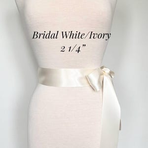 May include: Ivory satin bridal sash, 2 1/4 inches wide, tied in a bow. The text "Bridal White/Ivory" is above the sash. The sash is displayed on a mannequin.