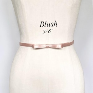 May include: A blush-colored belt with a bow detail. The belt is 3/8 inch wide and is displayed on a mannequin. The word "Blush" is printed above the belt.