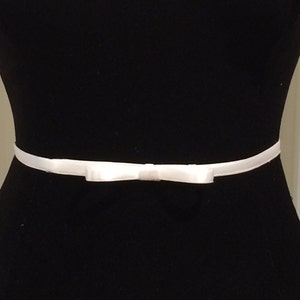 May include: A white satin ribbon belt with a small bow in the center. The belt is displayed on a black mannequin, highlighting the contrast between the bright white ribbon and the dark background. The bow is centered and adds a delicate touch.