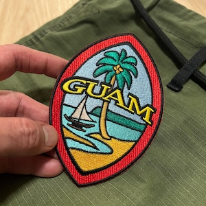May include: A red, white, and blue embroidered patch with the word "GUAM" in yellow lettering. The patch features a palm tree and a sailboat on a blue and green background.