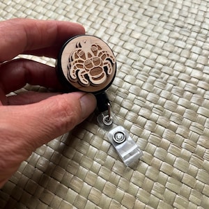 May include: A wooden badge reel with a laser-cut crab design. The crab is light brown and layered, set against a black reel. The reel has a clear plastic clip and a retractable cord. The design is detailed and artistic.