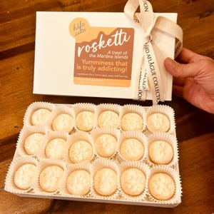 May include: A white box of Rosketti cookies from the Mariana Islands, with a ribbon tied around it. The box has text that reads "Yumminess that is truly addicting!" and the ingredients. The cookies are in individual paper cups.