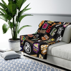 May include: Black throw blanket with a colorful pattern of Hawaiian-themed patches and text. The patches include phrases like "Island Girl", "Got Kelaguen", and "Land Style".