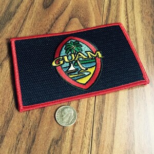 May include: A rectangular patch with a black background and a red border. The patch features an oval design with the word "GUAM" in yellow, a palm tree, and a beach scene. A dime is next to the patch.