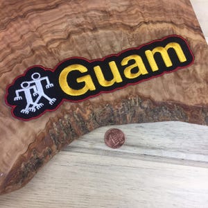 May include: Embroidered patch featuring the word "Guam" in yellow letters with a black border and red outline. Includes a white and black graphic of two figures. A penny is next to the patch.