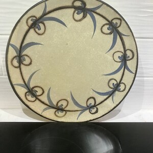 May include: A round ceramic plate with a cream-colored background and a blue and brown floral design around the edge.