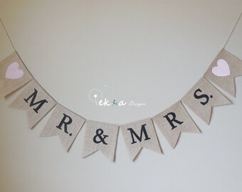 Könnte beinhalten: Ein Banner aus Jute mit den Worten "MR. & MRS." in schwarzen Buchstaben und zwei rosa Herzen. Das Banner ist an einer weißen Schnur befestigt.