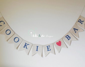 May include: A burlap banner with the words "Cookie Bar" in blue lettering and a red heart in the middle. The banner is strung on a white string and is hanging on a white wall.