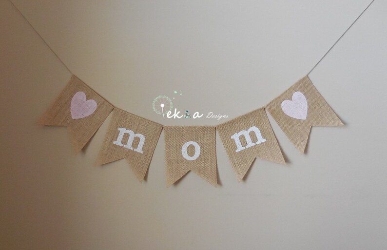 May include: A burlap banner with the word "MOM" spelled out in white letters. Each letter is on a separate flag, and there are pink hearts on the flags at either end of the banner.